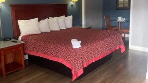Una cama bien hecha con una colcha estampada, una mesita de noche de madera, una lámpara y una silla en una habitación con paredes azules y suelo de madera.
