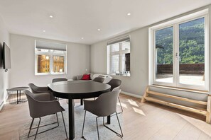 A modern room with a round table, six chairs, a TV, and large windows with a view of a forested area.