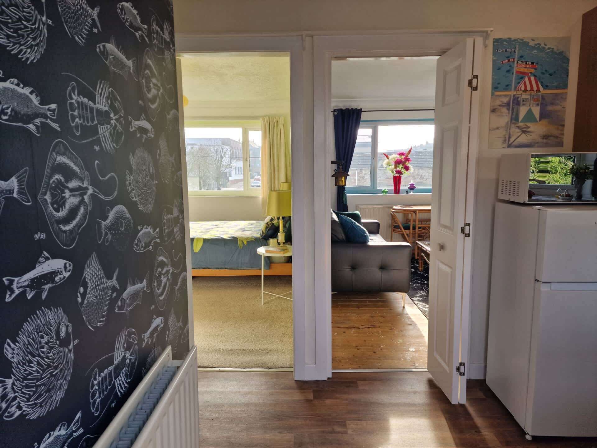 A hallway leading to a bedroom with a bed, a dining area with a table and chairs, and a kitchenette with a refrigerator.