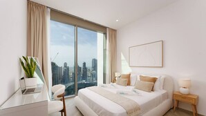 Ein modernes Schlafzimmer mit einem großen Bett, einem Schreibtisch, einem Stuhl, einem Nachttisch, einer Lampe und Blick auf eine Stadtlandschaft durch große Fenster.