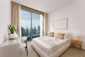 A modern bedroom with a large bed, a desk, a chair, a bedside table, a lamp, and a view of a cityscape through large windows. - Stunning 1 Bedroom Burj Khalifa View (Dubai)
