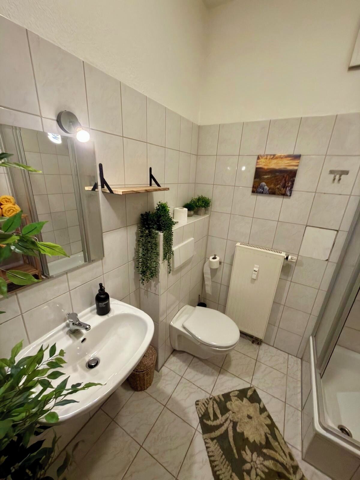 A bathroom with a sink, toilet, and shower area. There are plants on the shelves, a radiator, and a mirror.