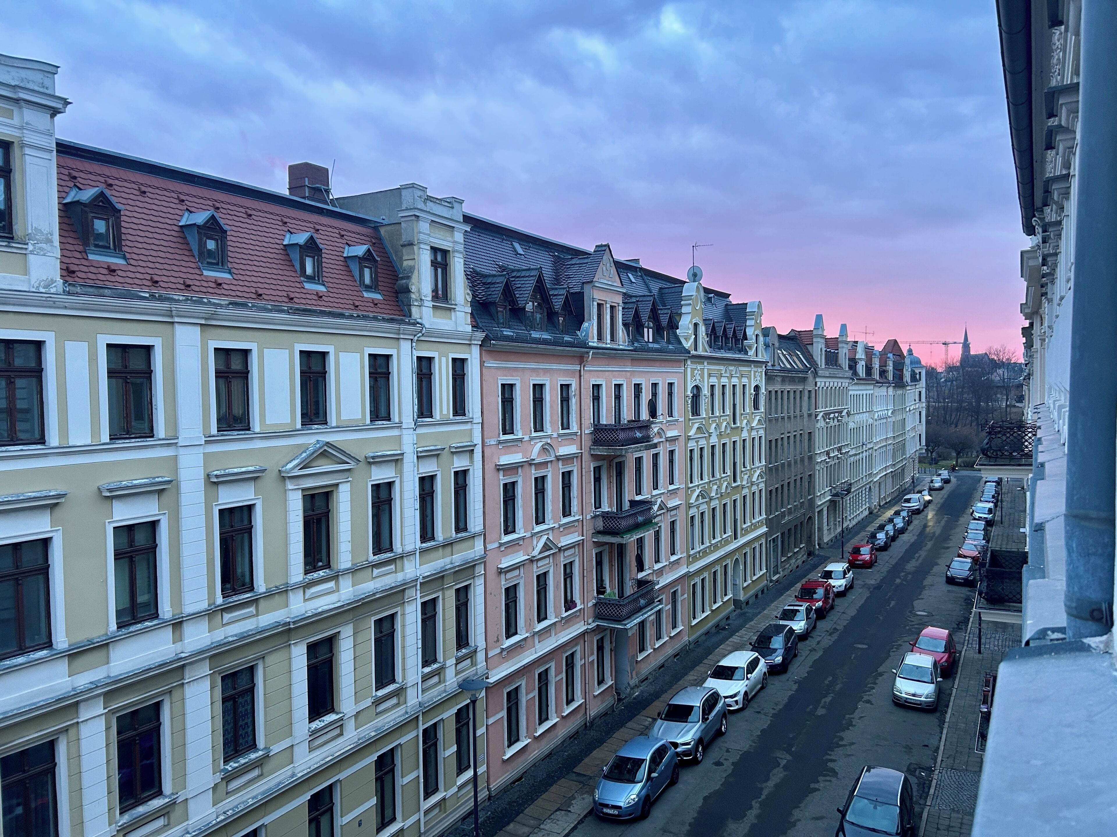 Une rangée d’immeubles à étages, tous de style architectural uniforme, une rue bordée de voitures garées et un ciel dégagé.