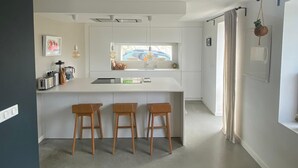 Una cocina moderna con una isla central, taburetes de bar, iluminación colgante y vista a un coche a través del ventanal.