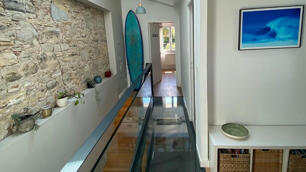 Un interior moderno con pared de piedra, barandilla de cristal, una obra de arte azul y vista a otro ambiente.