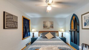 A bedroom with a bed, two bedside tables with lamps, a ceiling fan, artwork on the walls, and a door labeled 'ROME'.