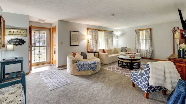 A living room with a sofa, ottoman, and a coffee table. There is a door with a sign, a lamp, and a mirror. - 1 Mi to Dtwn Tiffin! Brick Bungalow w/ Patio, Yard (Tiffin)