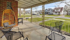 A covered porch with wicker chairs, a metal table, and a view of a residential street.