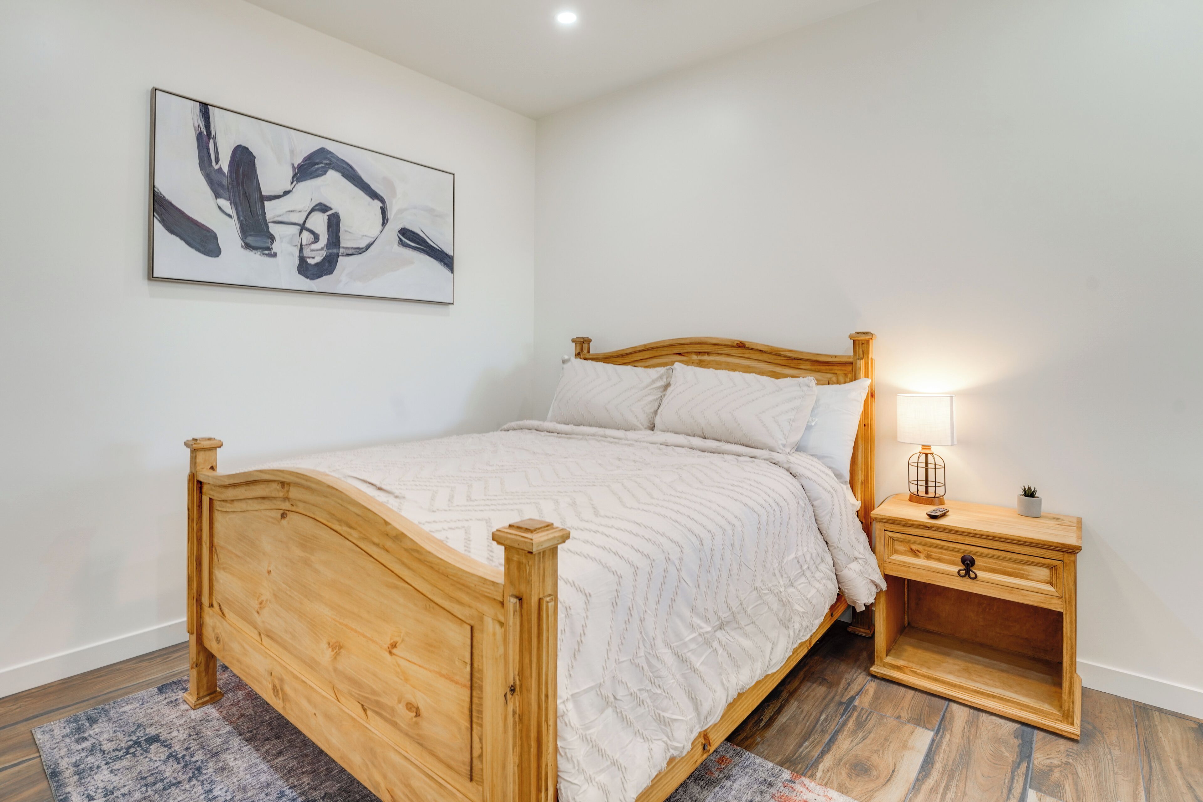 A bedroom with a wooden bed, a nightstand with a lamp, and a wall-mounted abstract painting.