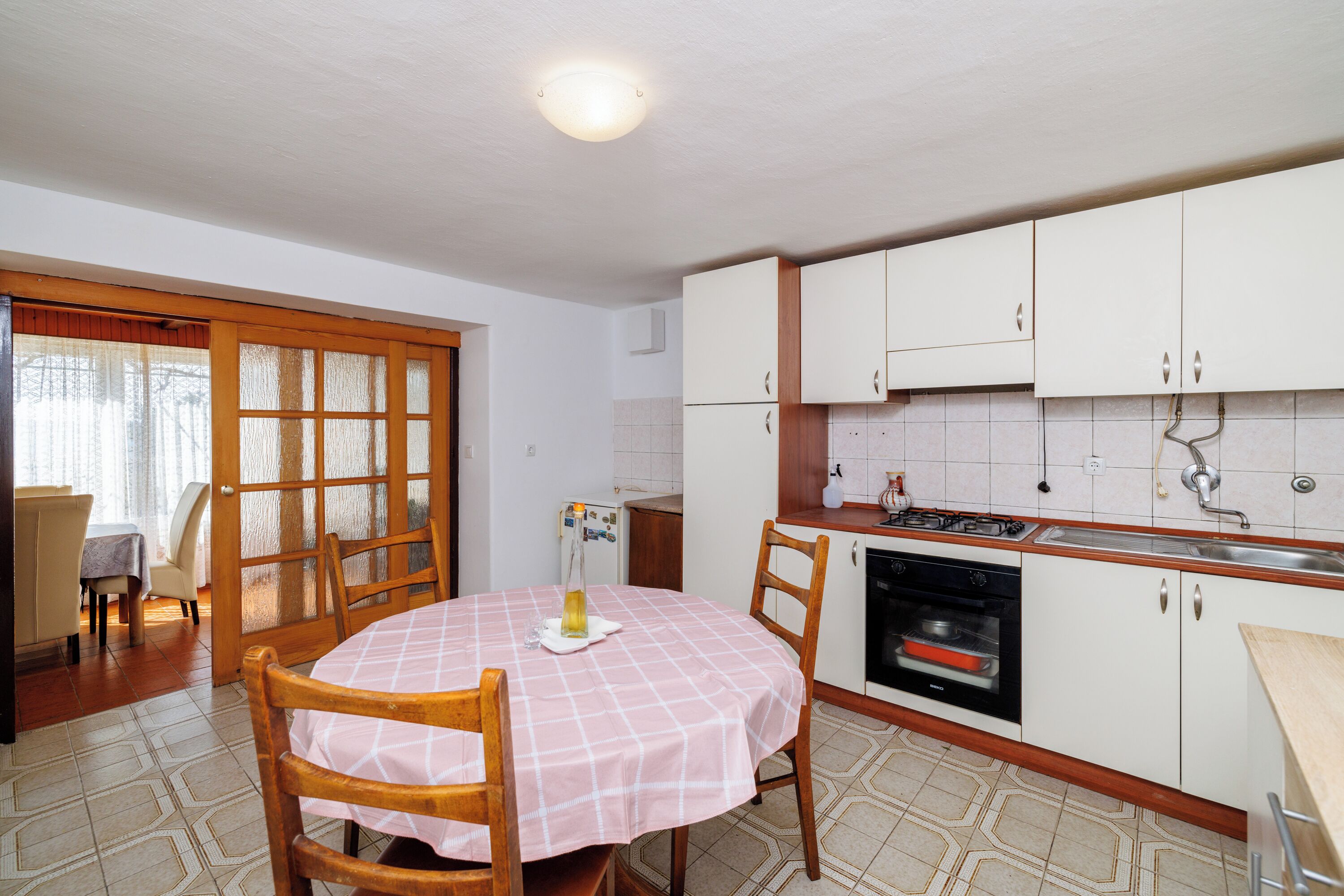 A dining area with a table set for two, a kitchen with white cabinets and a stove, and a door leading to an adjacent room.