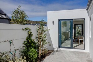 Ein modernes Haus mit weißer Fassade, eine Glasdoor die zu einer Terrasse mit Tisch und Stühlen führt, und ein kleiner Gartenbereich mit Pflanzen und einem Zaun.