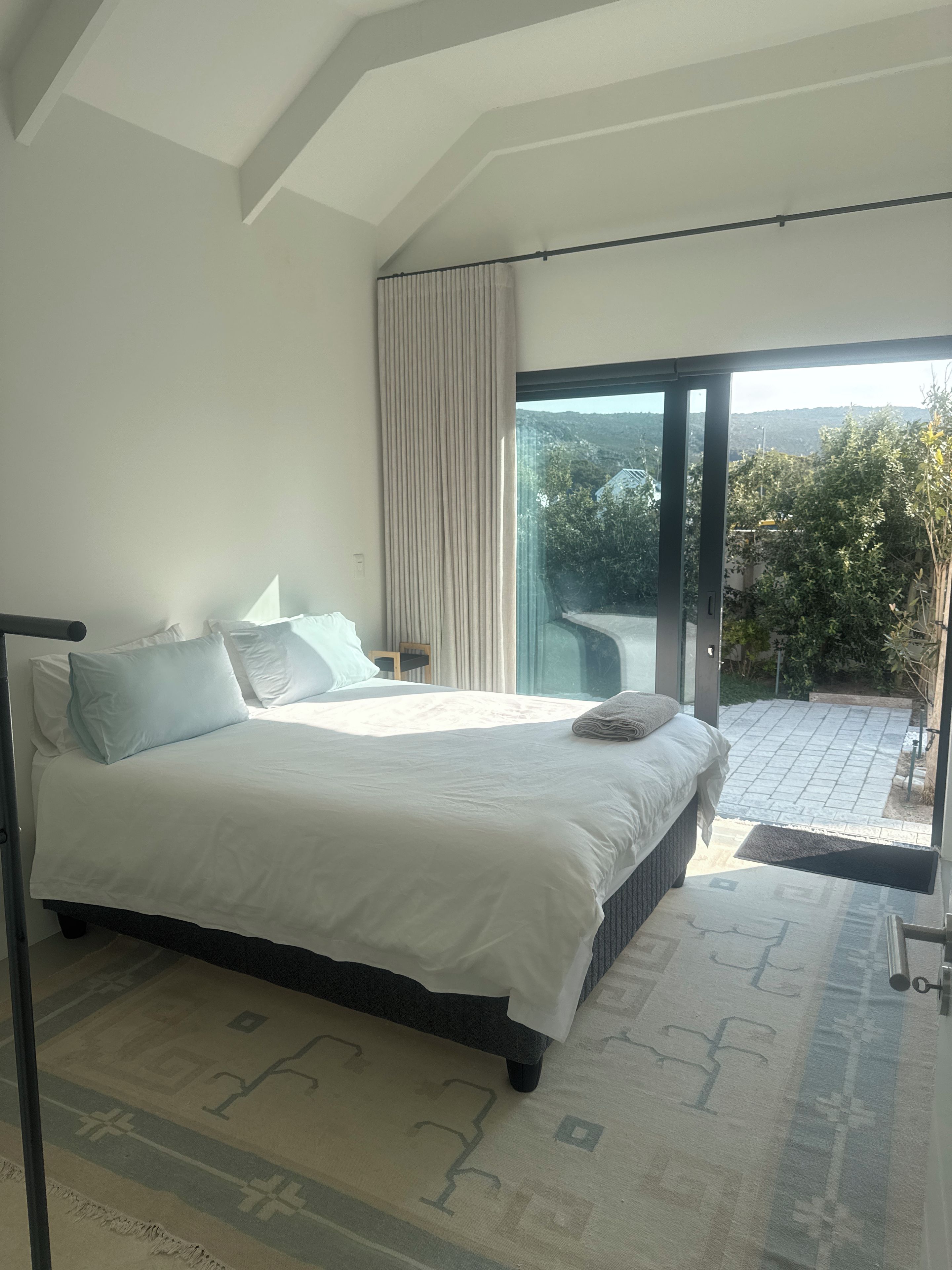 Un dormitorio con una cama grande, una mesita de noche, una silla y vistas a un jardín a través de una puerta corredera de vidrio.