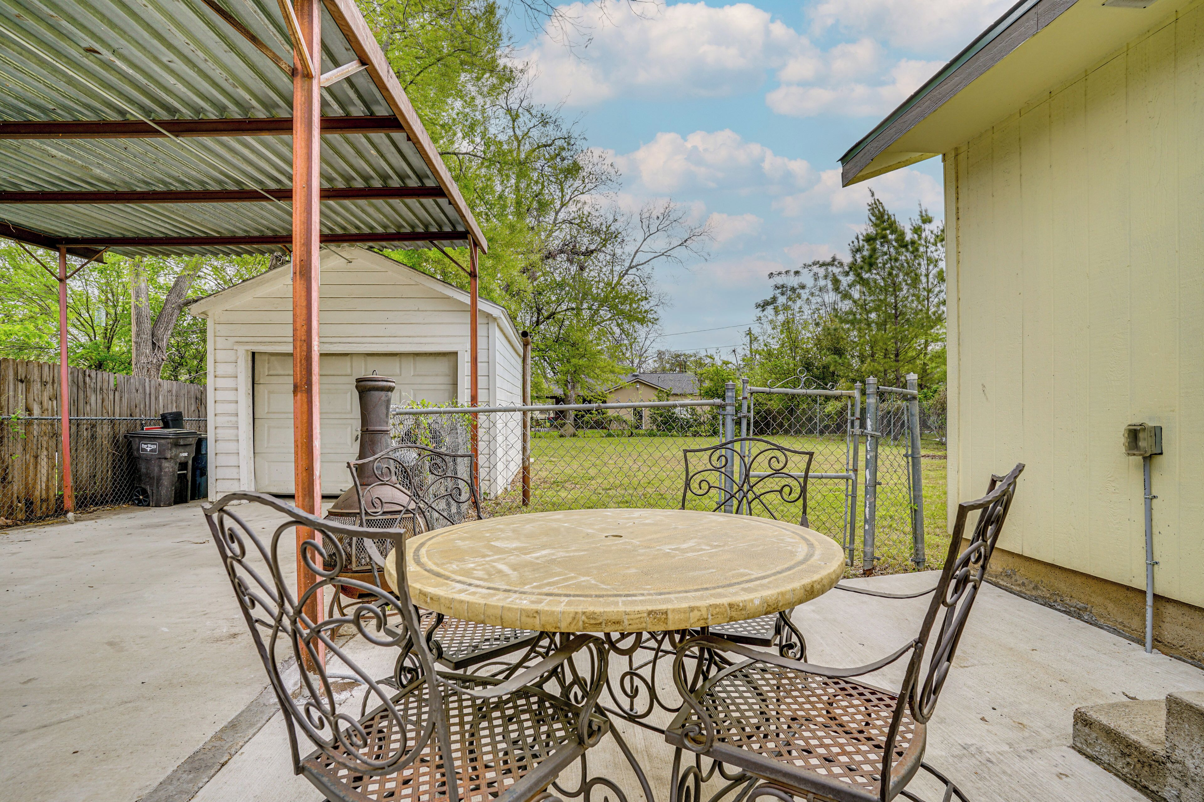 A patio with a round table and metal chairs, a covered area, and a small shed in the background.