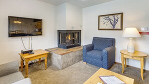 Un salon avec une cheminée, un téléviseur fixé au mur, un fauteuil à motifs, une petite table et une lampe sur une table d’appoint.