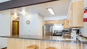 A kitchen with wooden cabinets, a stainless steel refrigerator, a microwave, and a coffee maker. There are three wooden stools at the counter. - Condo 3 Mi to Winter Park Resort: Community Perks! (Winter Park)