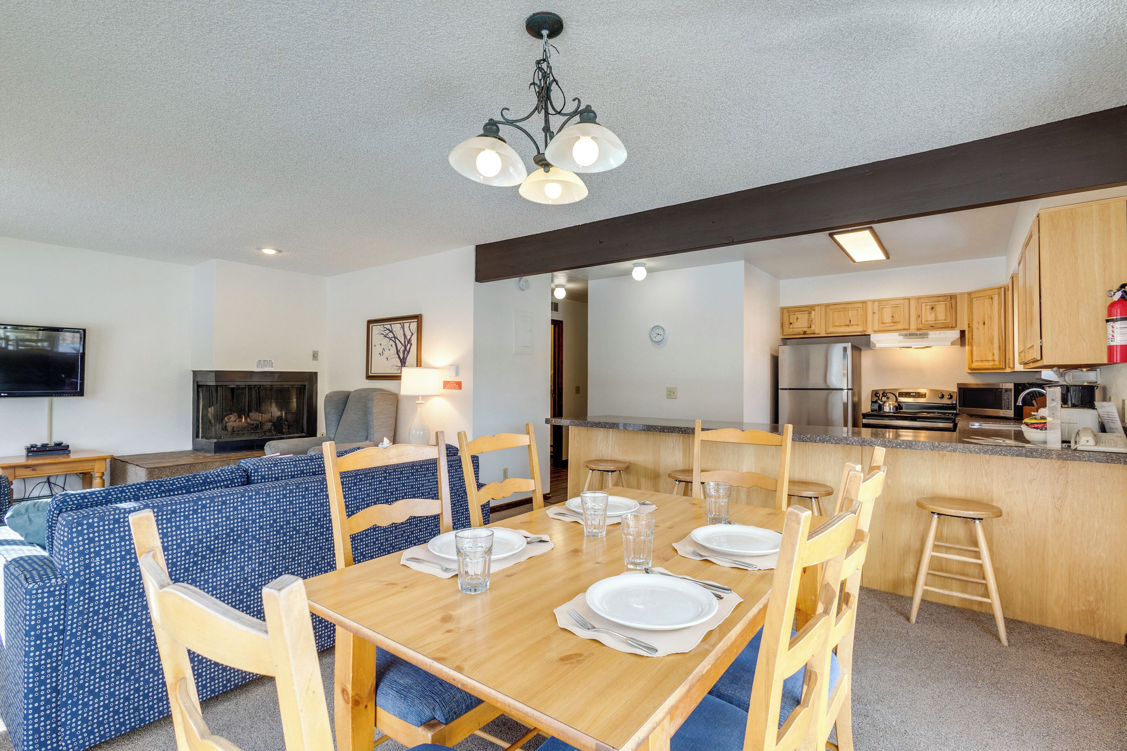 A dining area with a wooden table set for four, blue upholstered chairs, a kitchen with wooden cabinets, and a television in the living area.