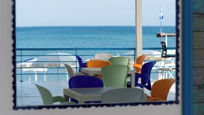 Une vue à travers une fenêtre donnant sur un espace de restauration avec des tables et des chaises, et offrant un panorama sur la mer.