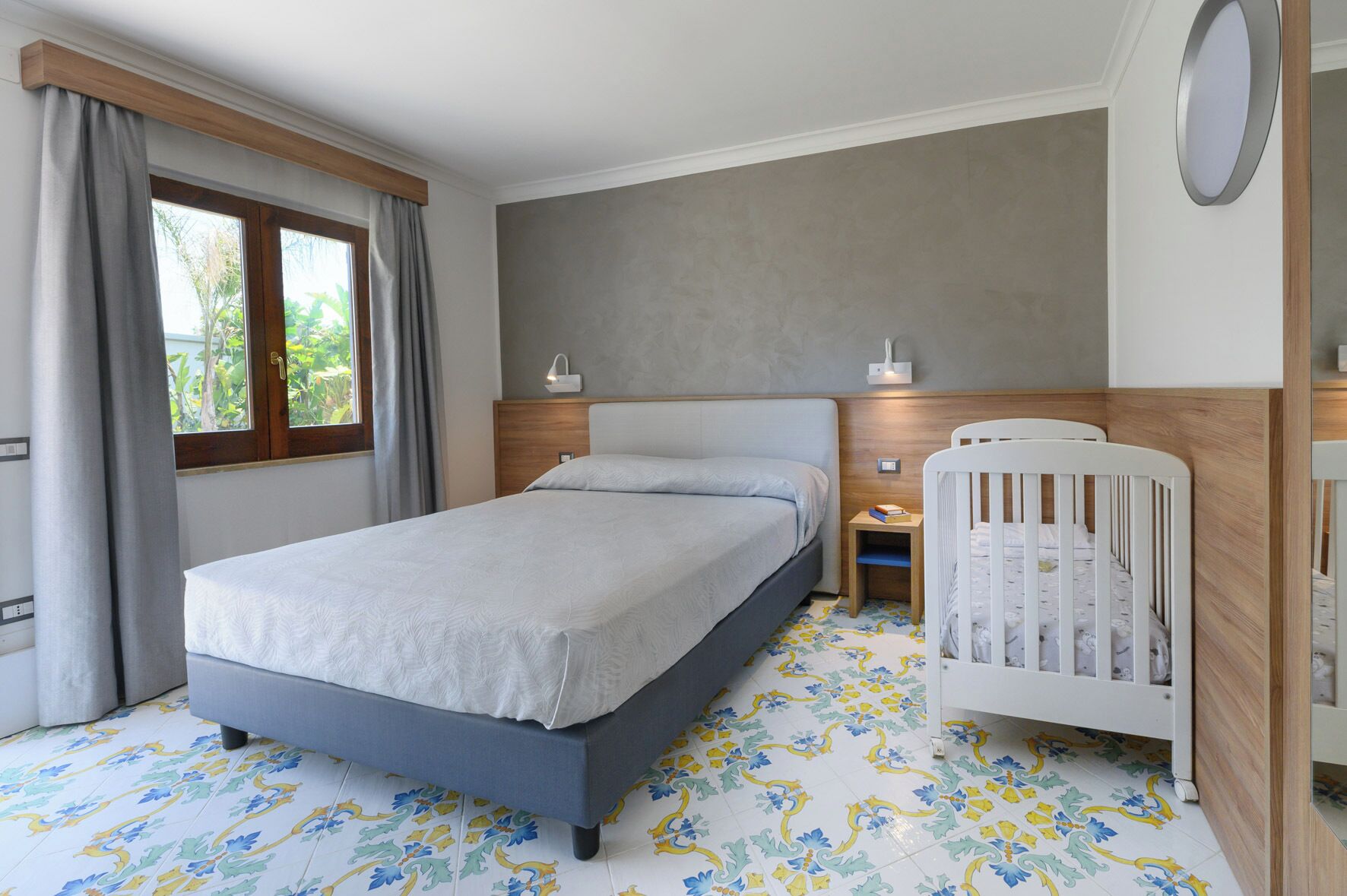 Un dormitorio con cama, cuna, mesita de noche, lámpara de pared y ventana con cortinas.