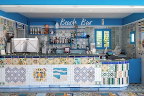 Un bar avec un mur recouvert de carreaux bleus et blancs derrière le comptoir, un comptoir où sont exposées différentes bouteilles, et une enseigne indiquant « Bar de plage ».