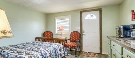 A bedroom with a bed, a chair, a desk, a lamp, a door, and a kitchenette with a microwave.