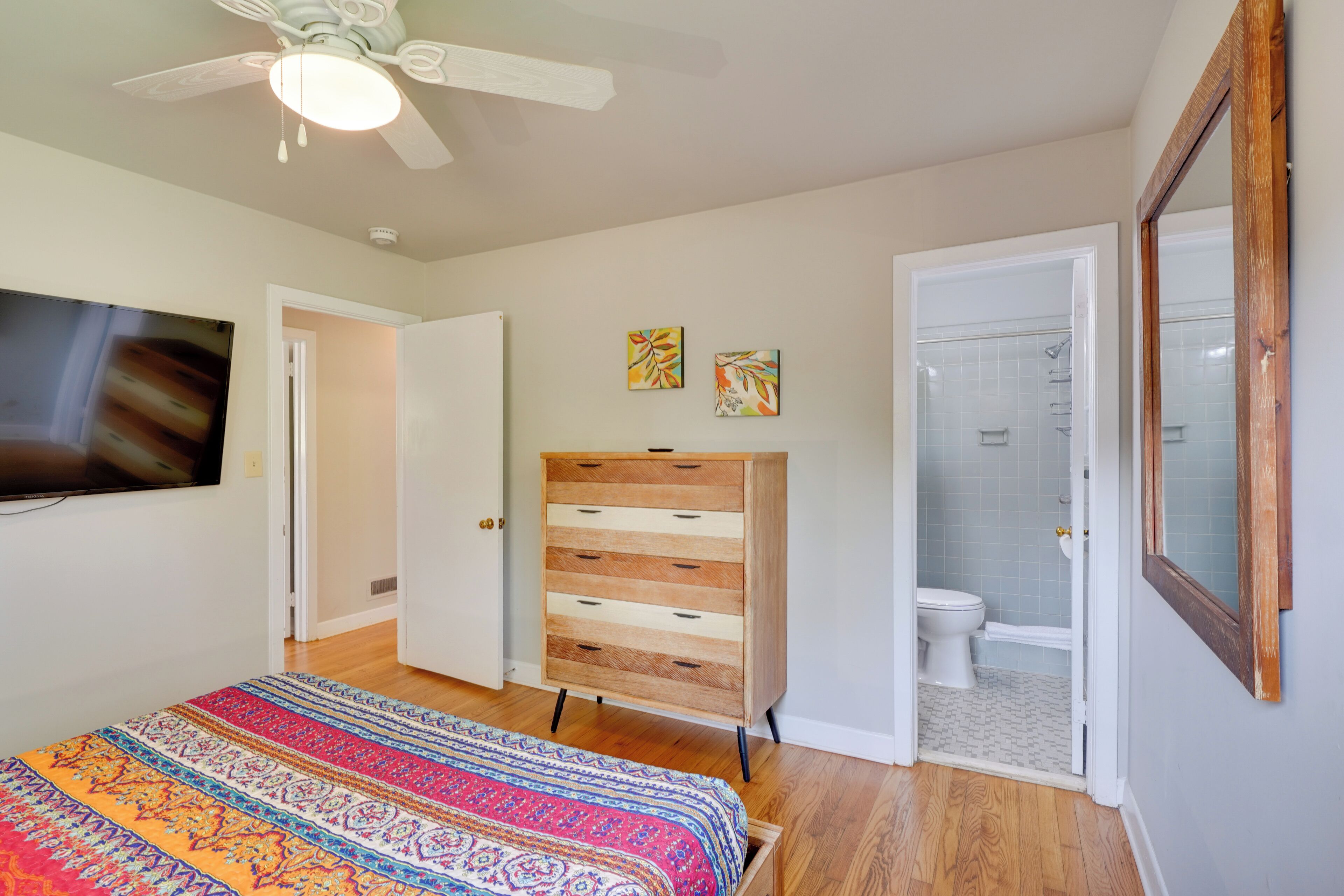 A bedroom with a bed, dresser, TV, ceiling fan, and a bathroom with a shower and toilet.