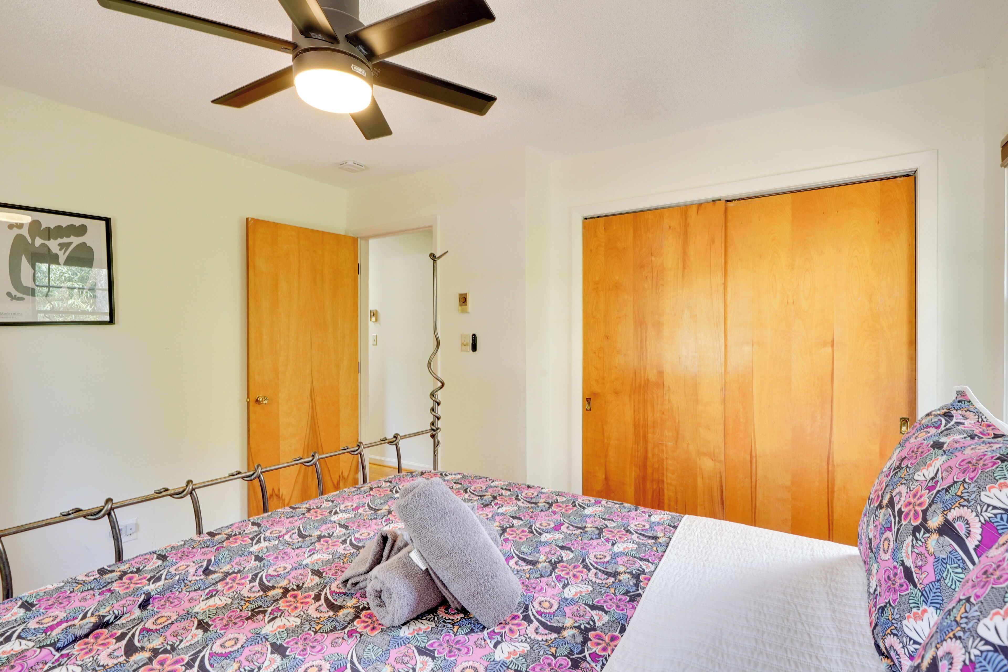 A bedroom with a bed, a ceiling fan, wooden doors, and a framed picture on the wall.