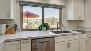 A modern kitchen with white cabinets, a stainless steel dishwasher, a sink, and a window with a view of the city. - Hot Tub, Pool & Socal Sunsets: San Diego Area Home (Spring Valley)