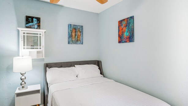 Une chambre à coucher comprenant un lit, une table de chevet, une lampe, un ventilateur de plafond et trois tableaux accrochés au mur.