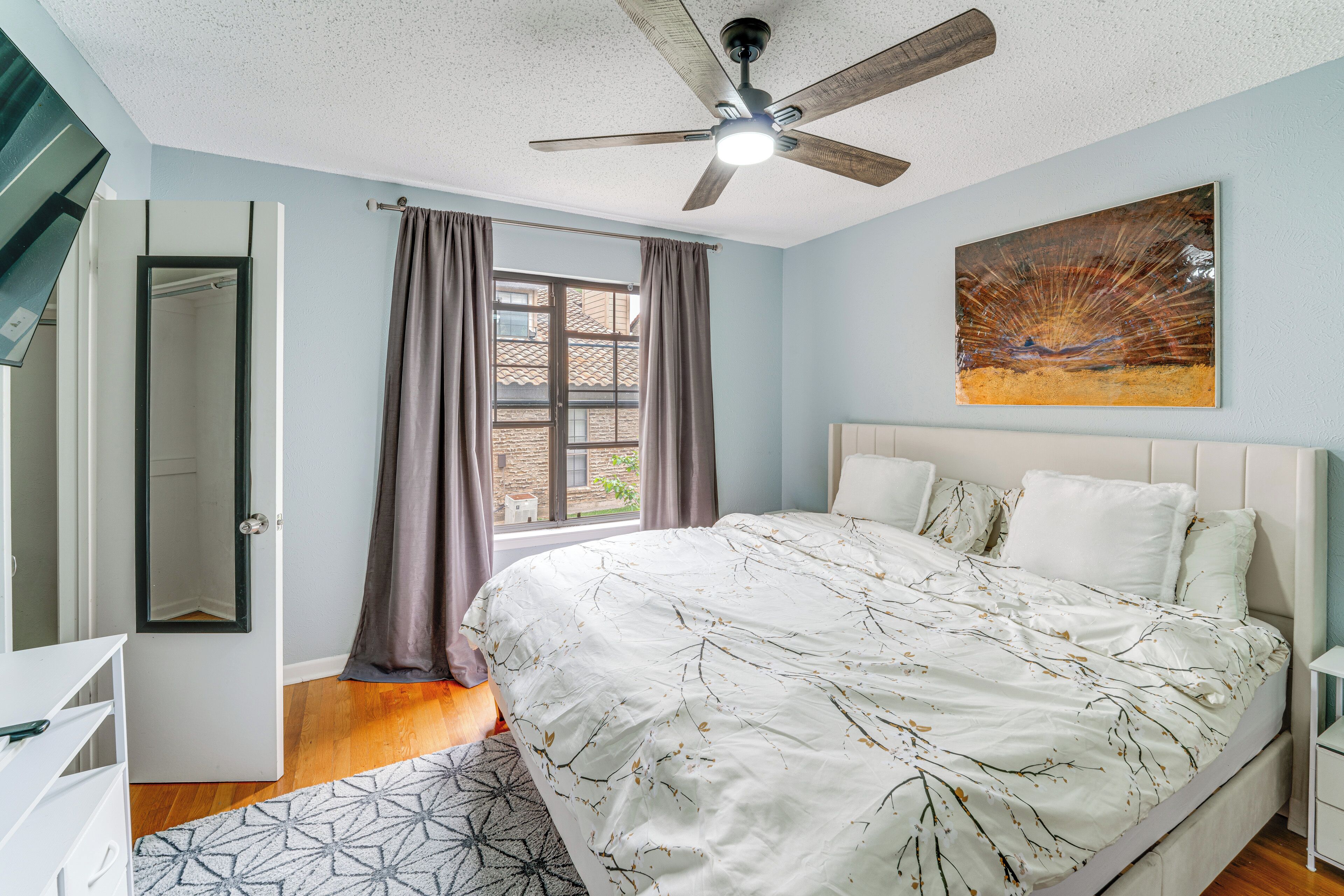 Une chambre à coucher avec un lit, un ventilateur de plafond, une fenêtre avec des rideaux, un tableau au mur et une porte donnant sur une autre pièce.