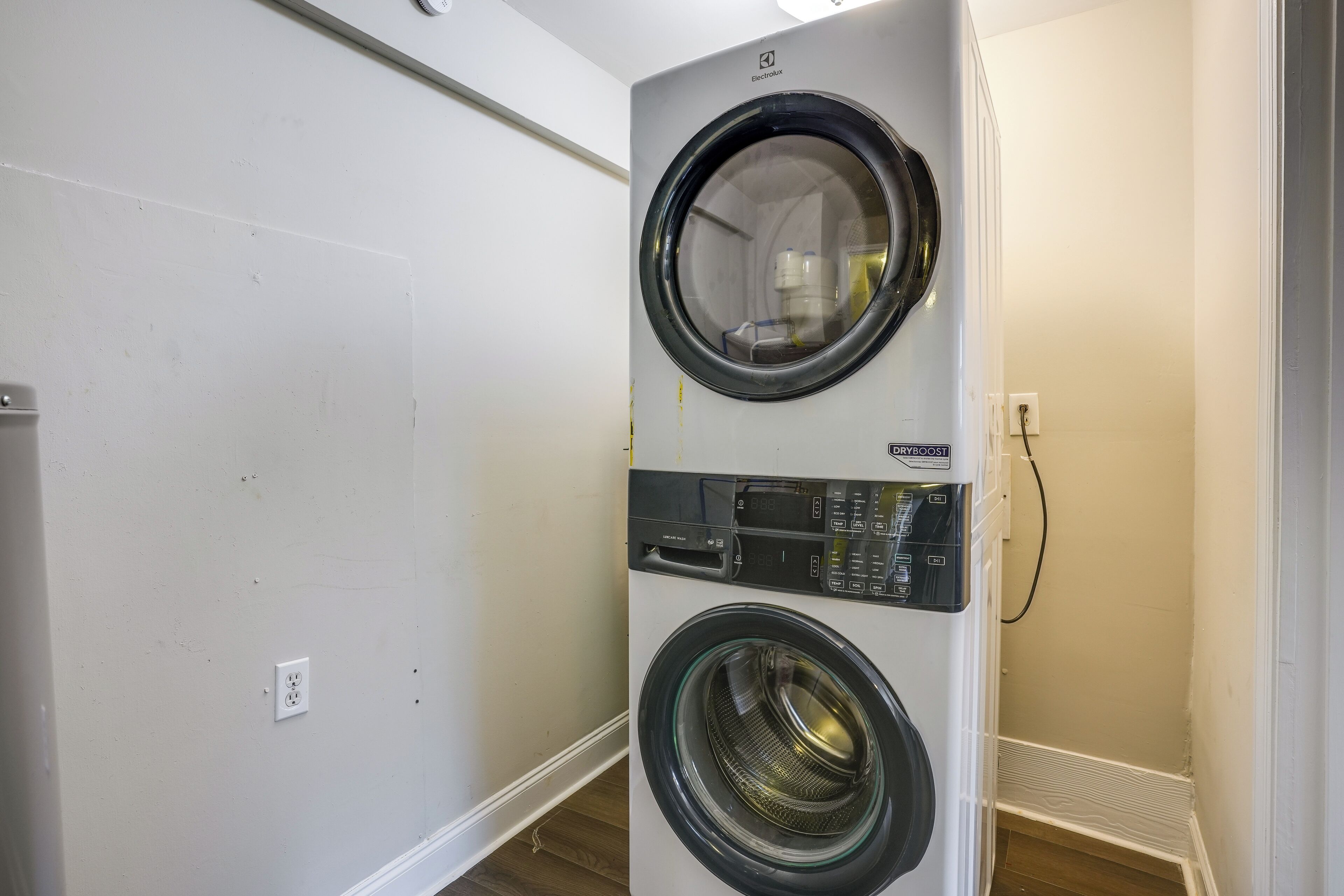 A wall-mounted front-loading washer and dryer unit in a laundry room with a plain wall and a wooden floor.