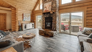 A log cabin interior with a stone fireplace, wooden walls, and a view through large windows. - Group Retreat in the Bitterroot Valley w/ Hot Tub (Victor)