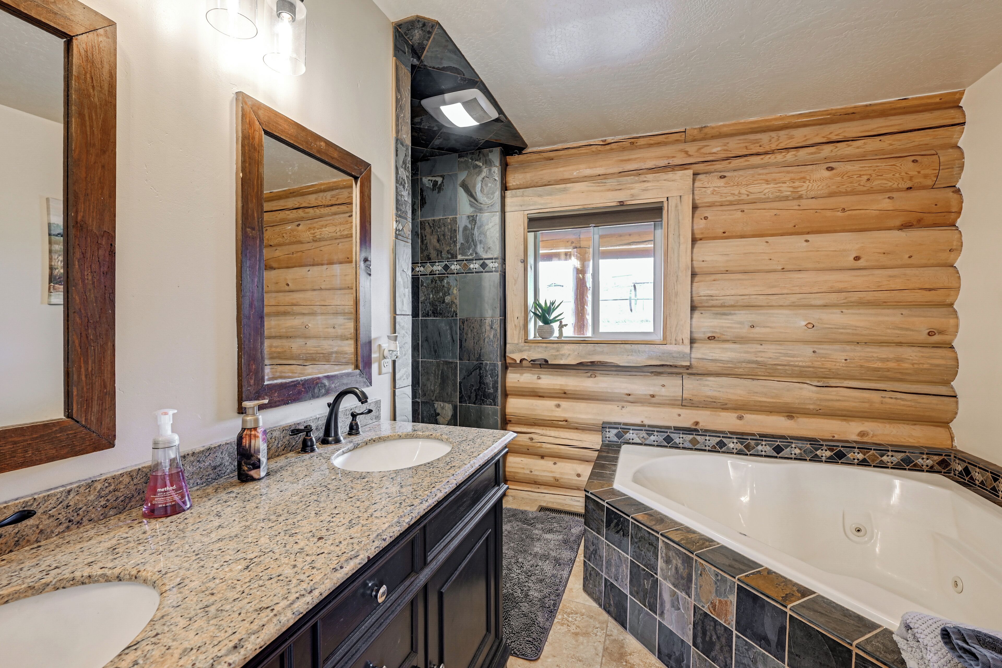 A bathroom with a wooden log wall, a large mirror, two sinks, a bathtub, and a window with a view.