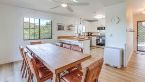 Una cocina moderna con armarios blancos, un comedor con mesa de madera y sillas de cuero, un ventilador de techo y vistas al exterior con vegetación.