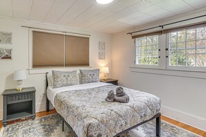 A bedroom with a bed, a nightstand with a lamp, a window with blinds, and a view of trees. - Half-mi to Dtwn Wendell! 60s Farmhouse w/ Fire Pit (Wendell)