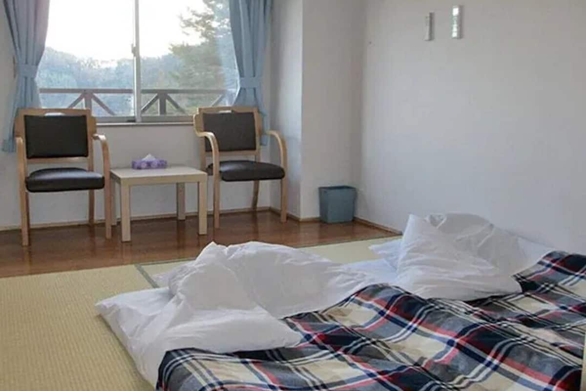 Ein Zimmer mit einem Bett, zwei Stühlen, einem kleinen Tisch, einem Fenster mit Vorhängen und einem Mülleimer.
