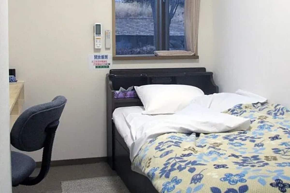 Ein kleines Schlafzimmer mit einem Bett, einem Stuhl, einem Fenster und einem Thermostat an der Wand.