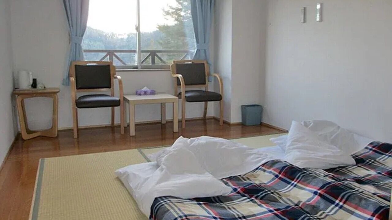 Una habitación con cama, dos sillas, una mesita y un ventanal.