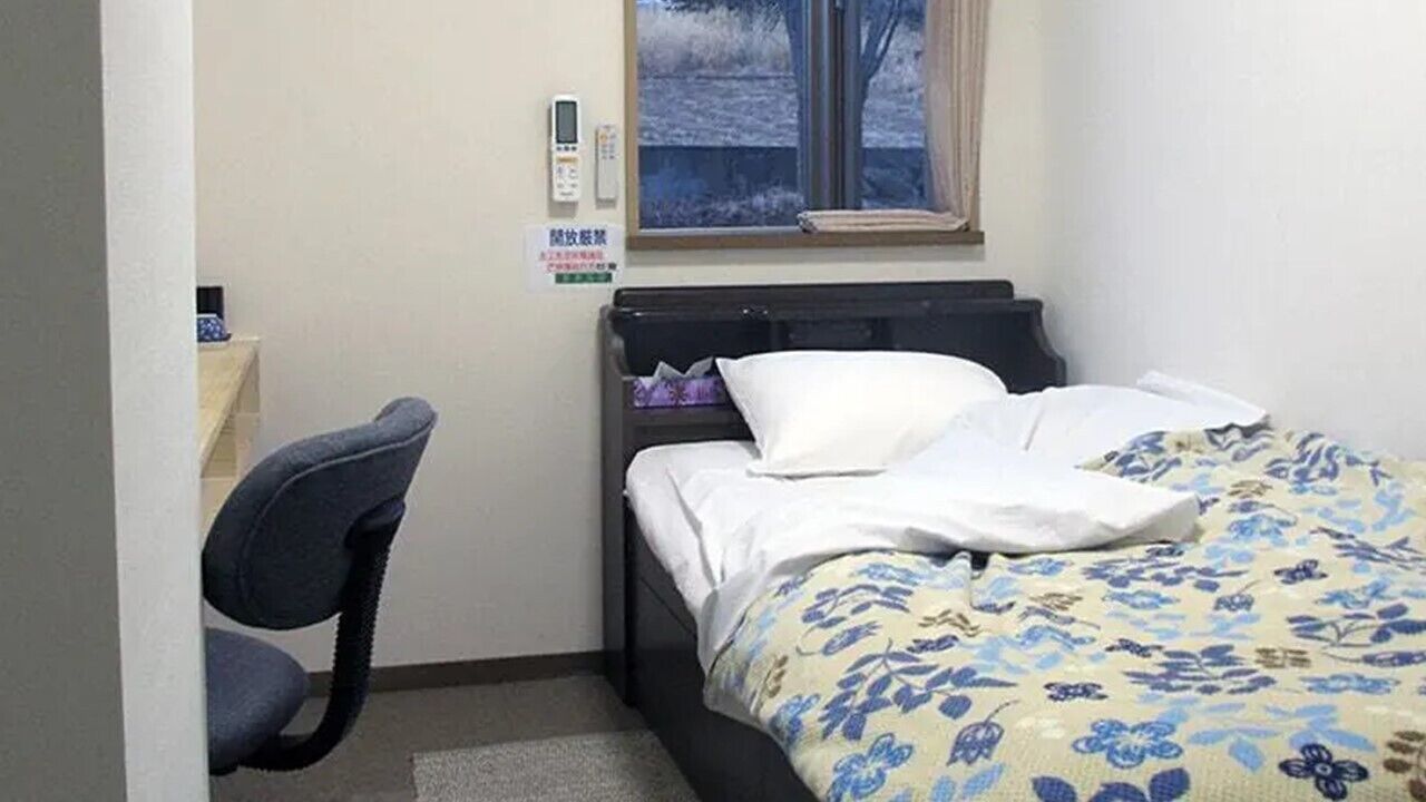 A small bedroom with a bed, a chair, a window, and a thermostat on the wall.