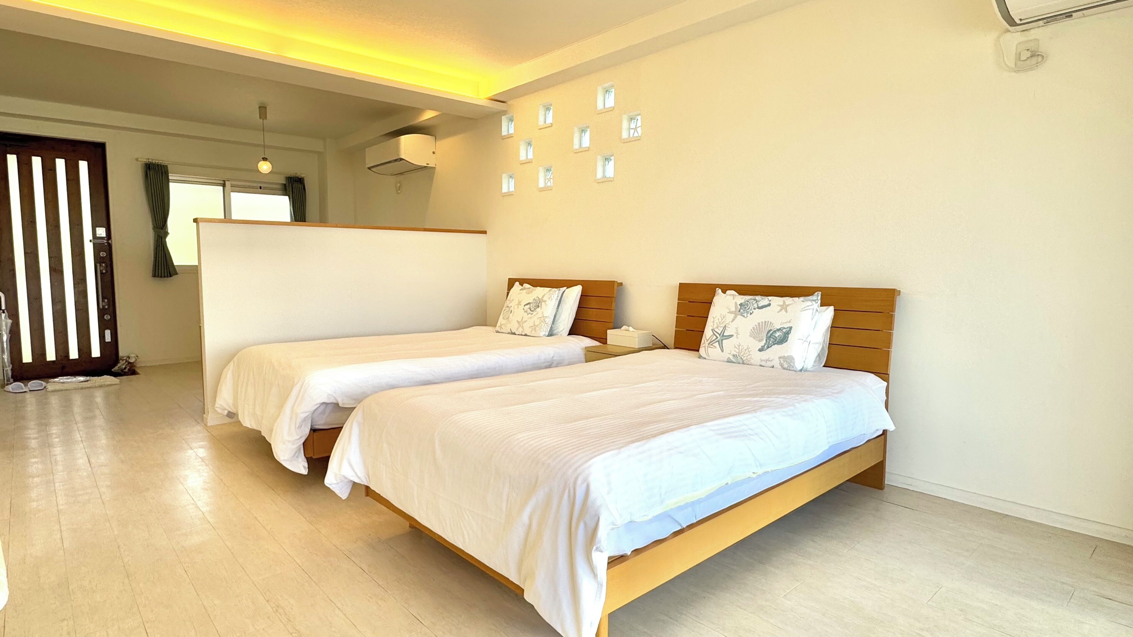 Habitación de hotel con dos camas individuales, cabecero de madera, una mesita de noche pequeña, aire acondicionado instalado en la pared y una puerta con persianas verticales.
