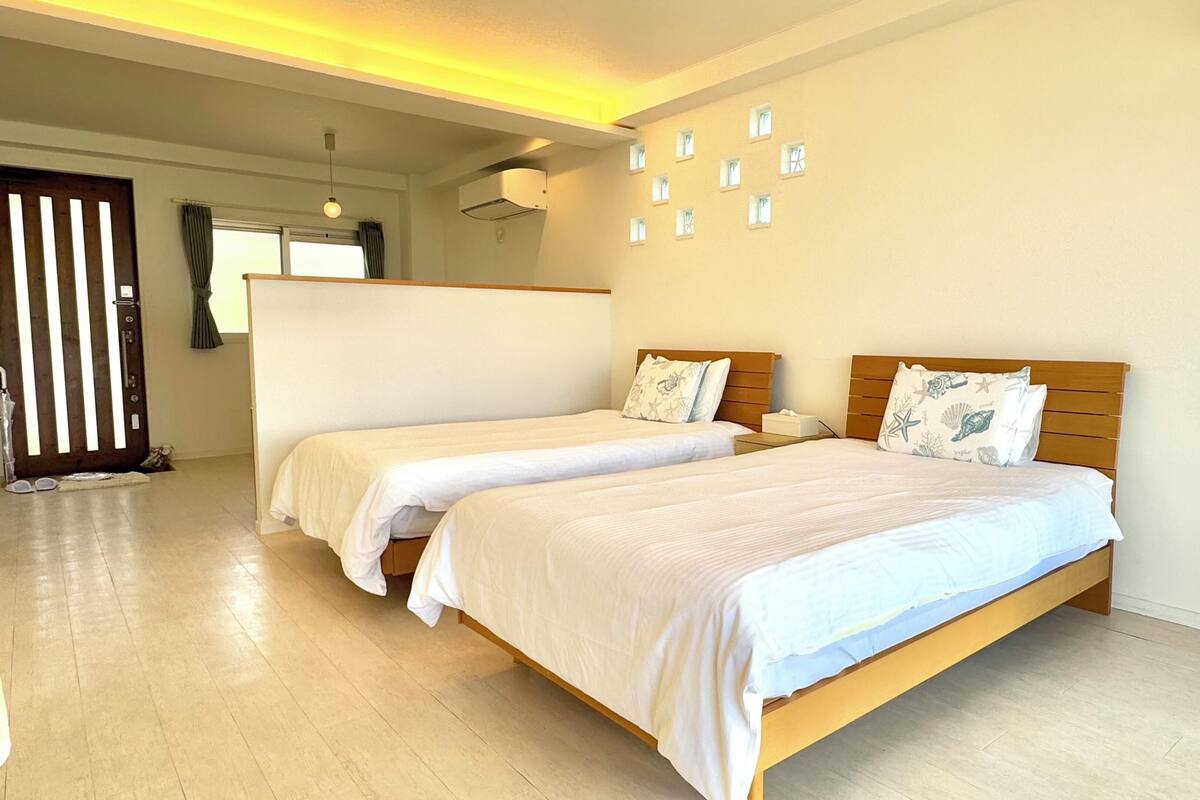 Habitación de hotel con dos camas individuales, cabecero de madera, una mesita de noche pequeña, aire acondicionado instalado en la pared y una puerta con persianas verticales.