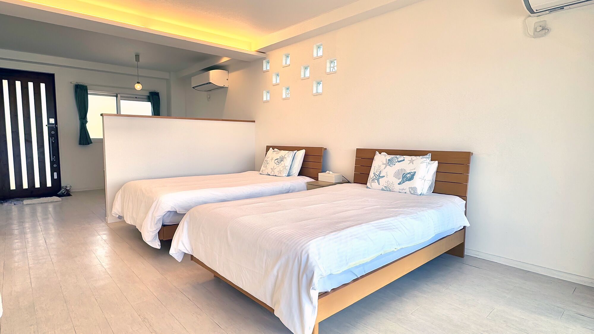 Habitación de hotel con dos camas individuales, cabecero de madera, una mesita de noche pequeña, aire acondicionado instalado en la pared y una puerta con persianas verticales.