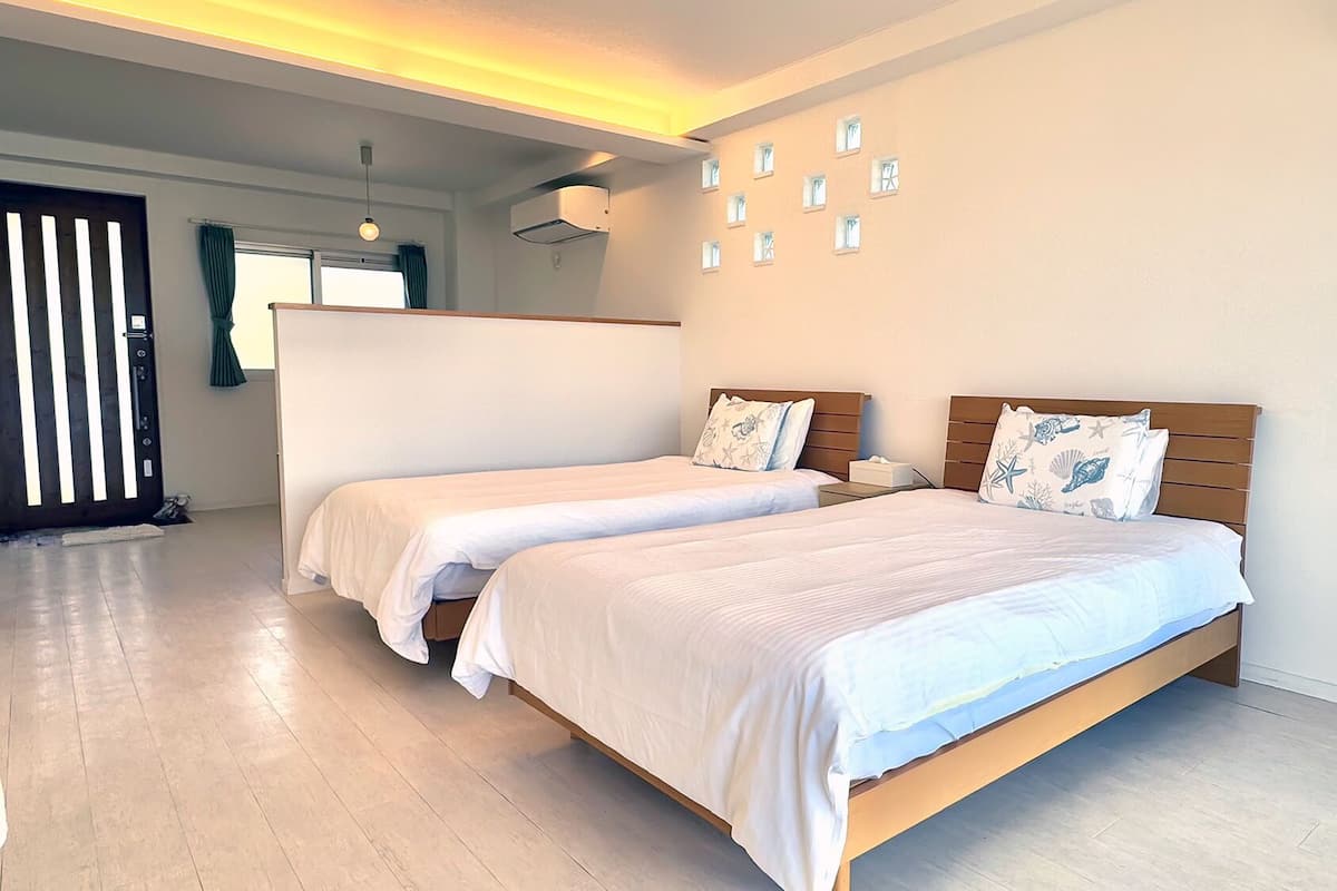 Habitación de hotel con dos camas individuales, cabecero de madera, una mesita de noche pequeña, aire acondicionado instalado en la pared y una puerta con persianas verticales.