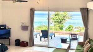 A room with a TV, a bed, a sofa, a table, a chair, a balcony with a view of the sea, and a ceiling fan.