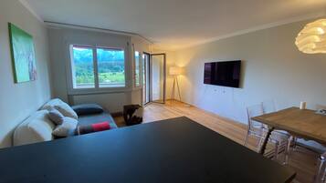 A modern living space with a sofa, a dining table, a TV, and a view of greenery through a large window.