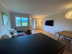 A modern living space with a sofa, a dining table, a TV, and a view of greenery through a large window.