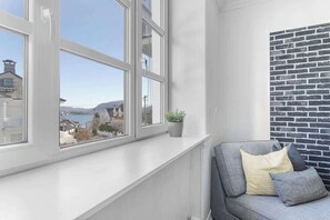 A room with a large window, a view of buildings and water, a grey sofa with cushions, and a brick-patterned wall.