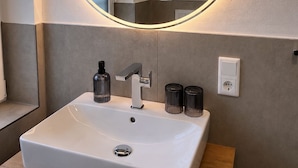A bathroom with a white sink, a mirror with a circular light, and a towel dispenser. - elf-36 (Esens)
