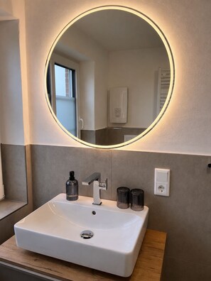 Une salle de bain avec un lavabo blanc, un miroir doté d’un éclairage circulaire et un distributeur de serviettes.