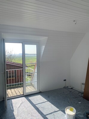 A room with a large sliding glass door leading to a balcony, a sloped ceiling, and a view of a distant landscape. - elf-36 (Esens)