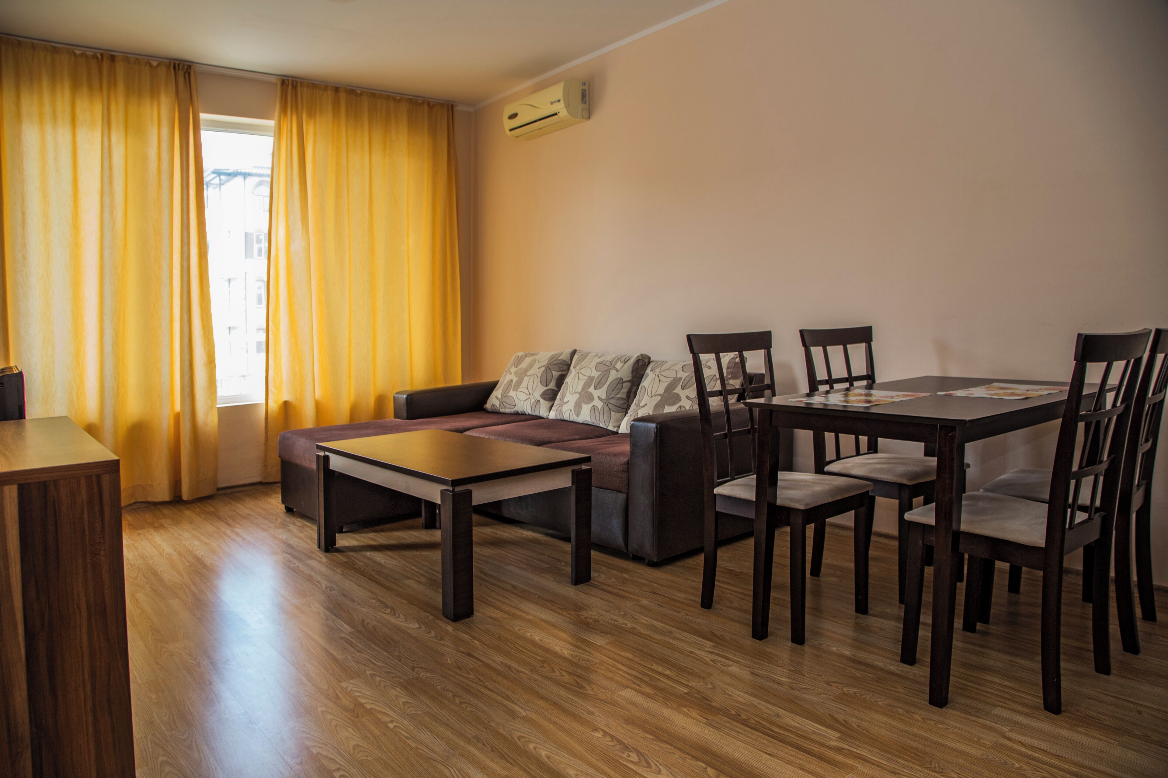 Ein Wohnzimmer mit einer Couch, einem Esstisch mit Stühlen, Holzboden und einem Fenster mit gelben Vorhängen.